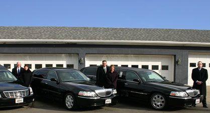 Five people in formal attire standing with black limousines in front of a garage.