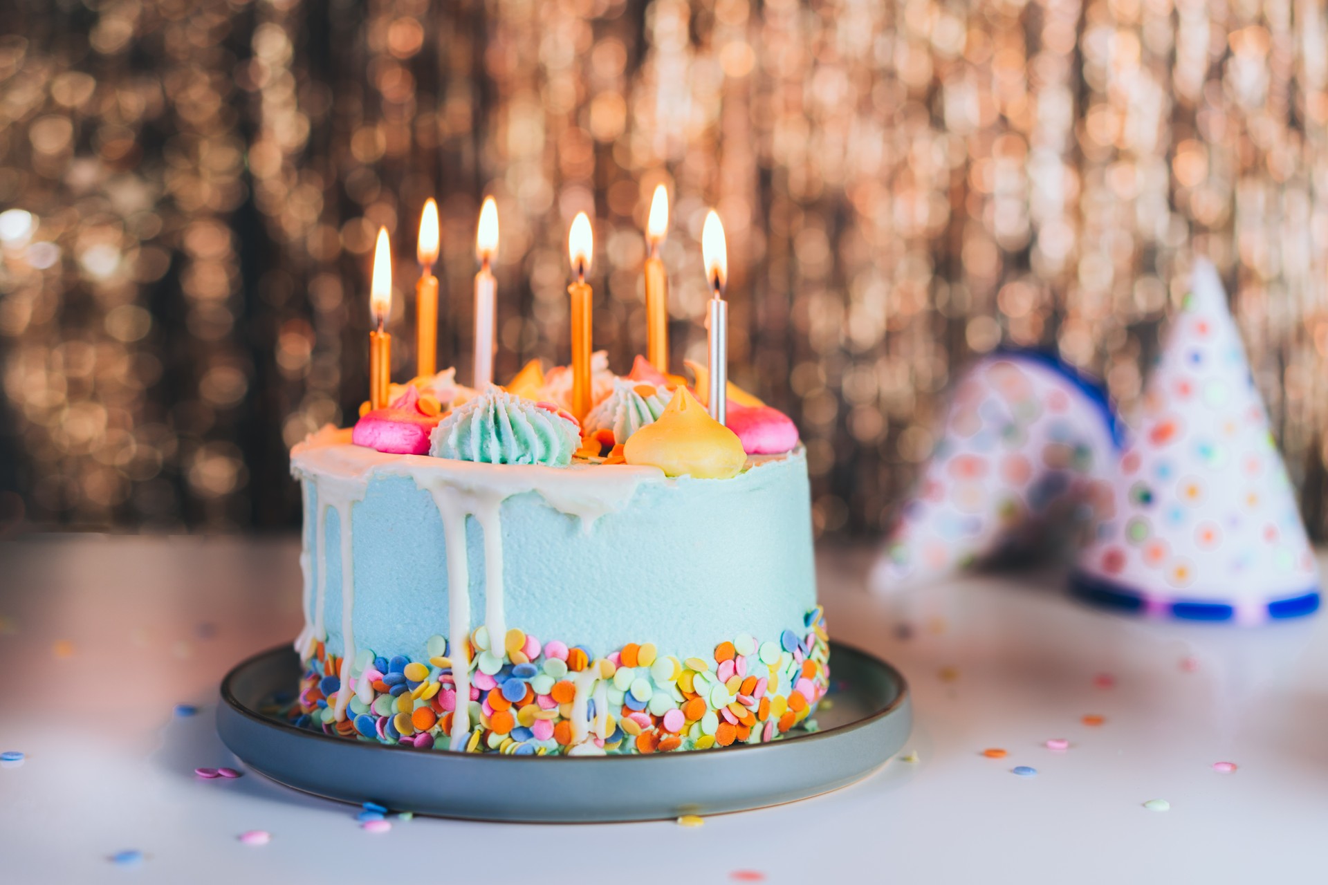 Colorful birthday cake with sprinkles and burning candles and festive caps on the sparkling gold tinsel background. Festive birthday celebration, party. Selective focus, copy space.